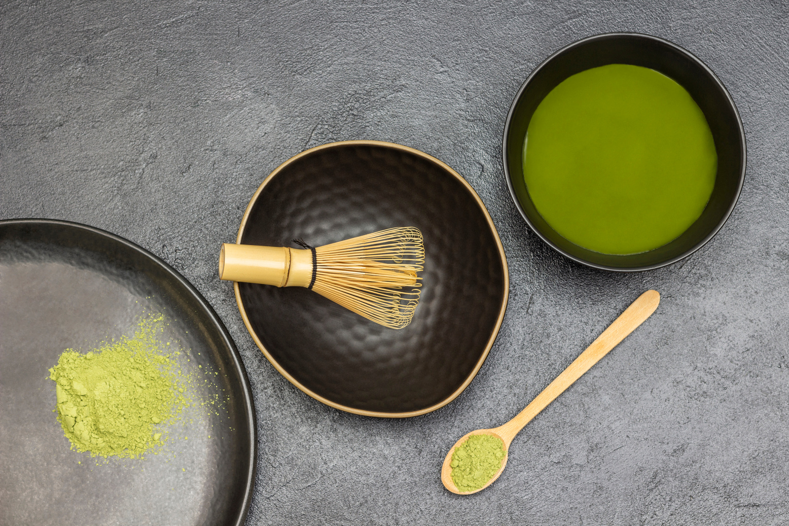 Three black bowls with green matcha powder and matcha tea. Wooden spoon with matcha powder